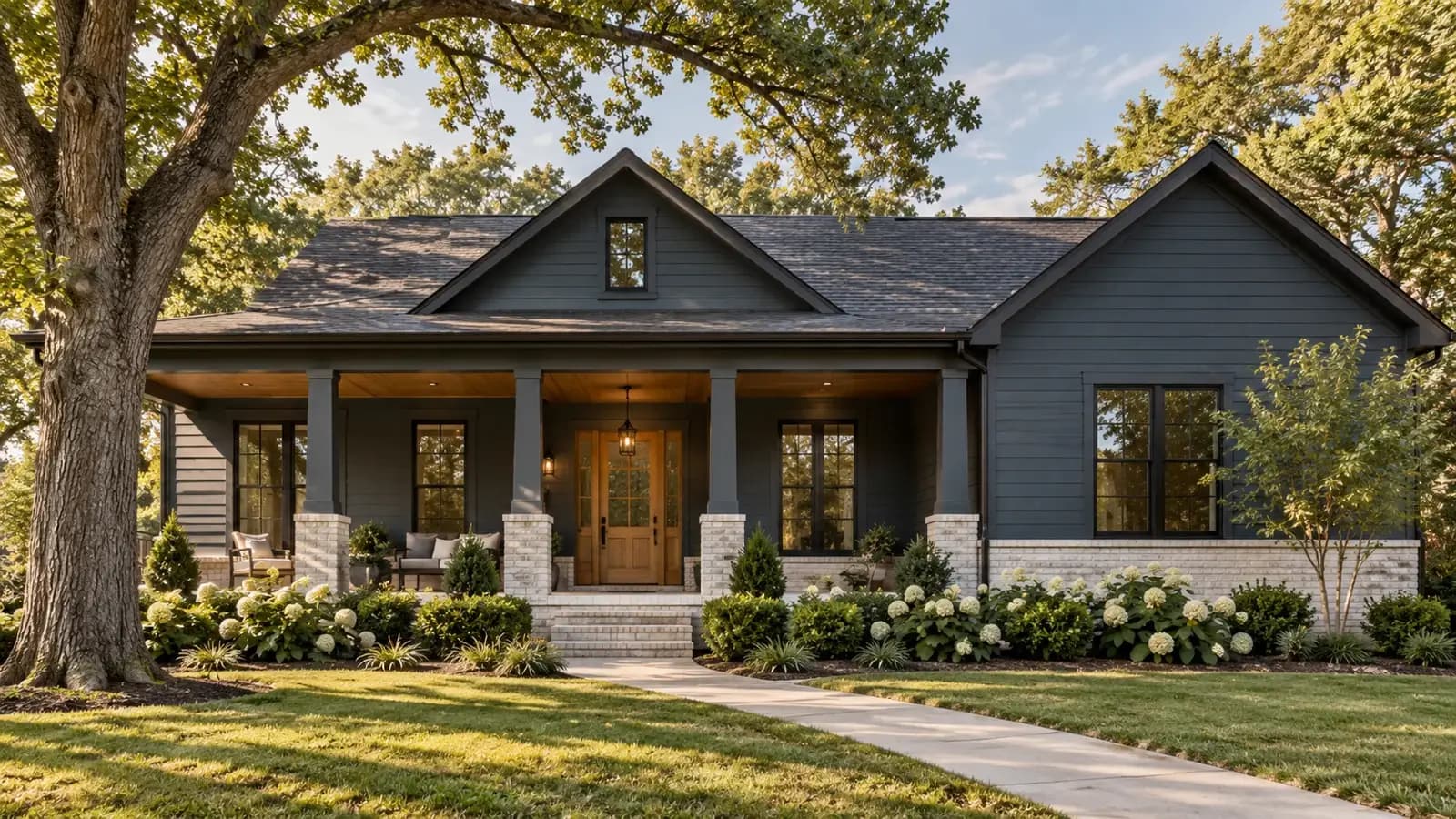 A grey-sided craftsman home with a dark shingle roof under mature oak trees